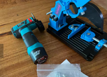 Top-down view of QuickMech angle grinder stand with installed cordless brushless grinder and red safety shield showing 0-45 degree adjustable bevel vise on cutting mat workbench
